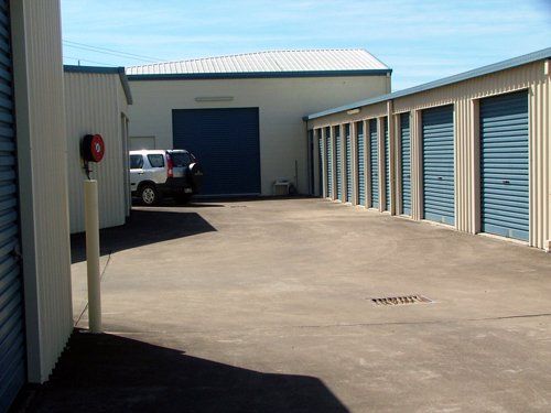 Storage units with blue doors and a large warehouse building. A car is parked in the paved area.