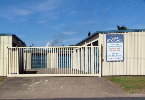 Closed self-storage facility gate with cream-colored units, sign, and blue sky. The gate is open.