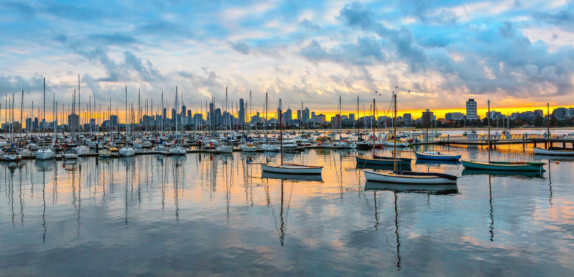 Boats docked in a harbor with a city skyline in the background, under a cloudy, colorful sky.