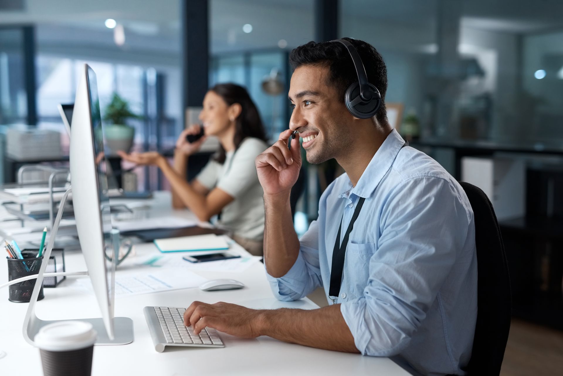 Man wearing a headset smiles while typing at a computer in a modern office. Another person is on the phone in the background.