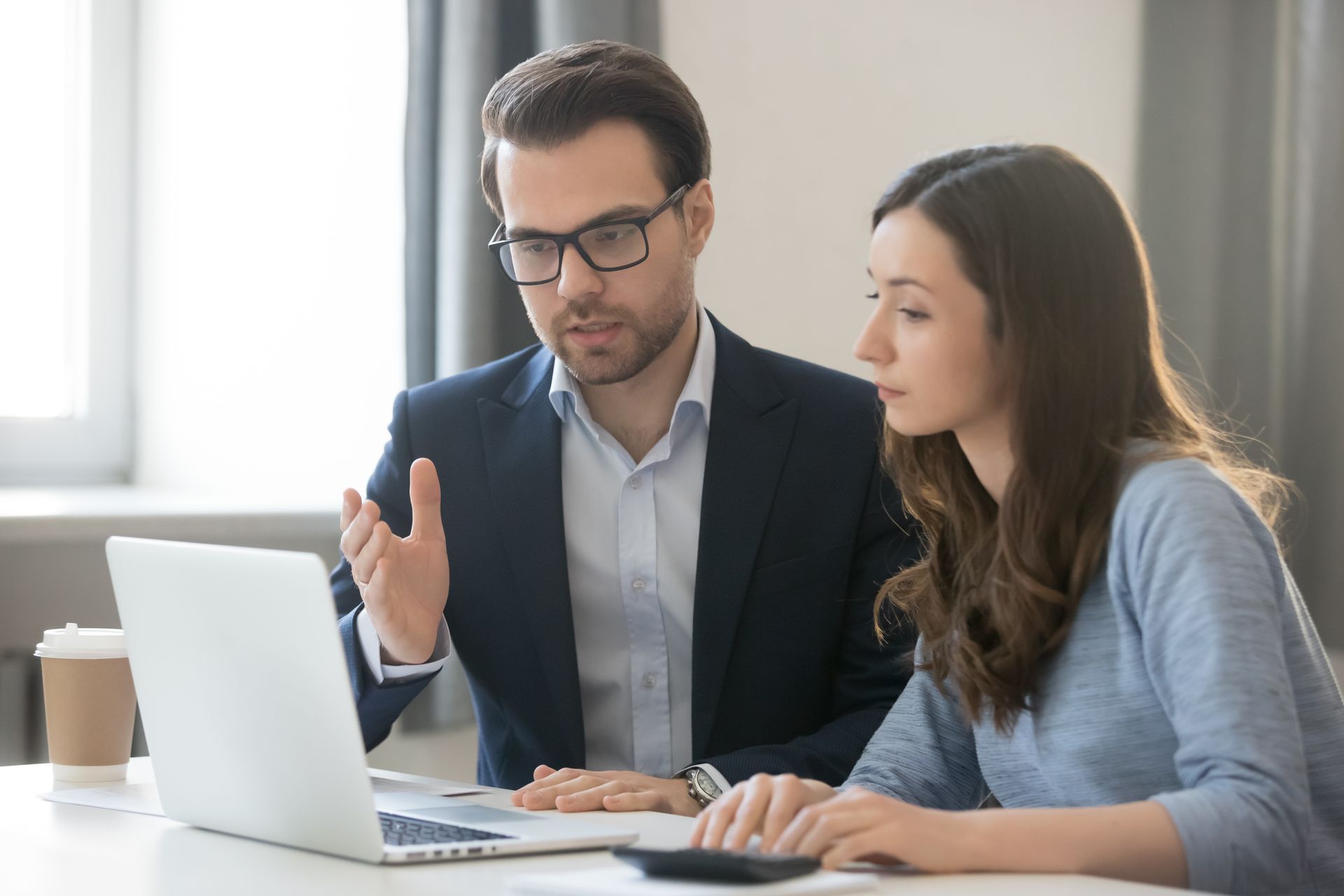 Man in suit gestures at laptop screen, explaining to woman in blue sweater.