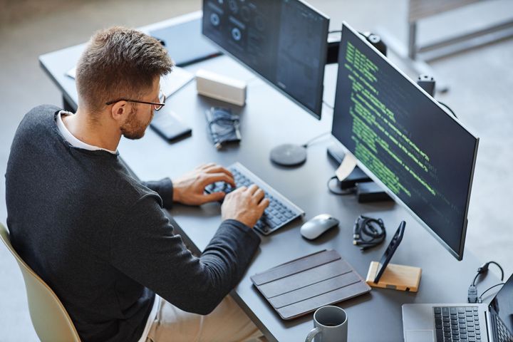 Person with glasses coding at a desk with two monitors displaying code.
