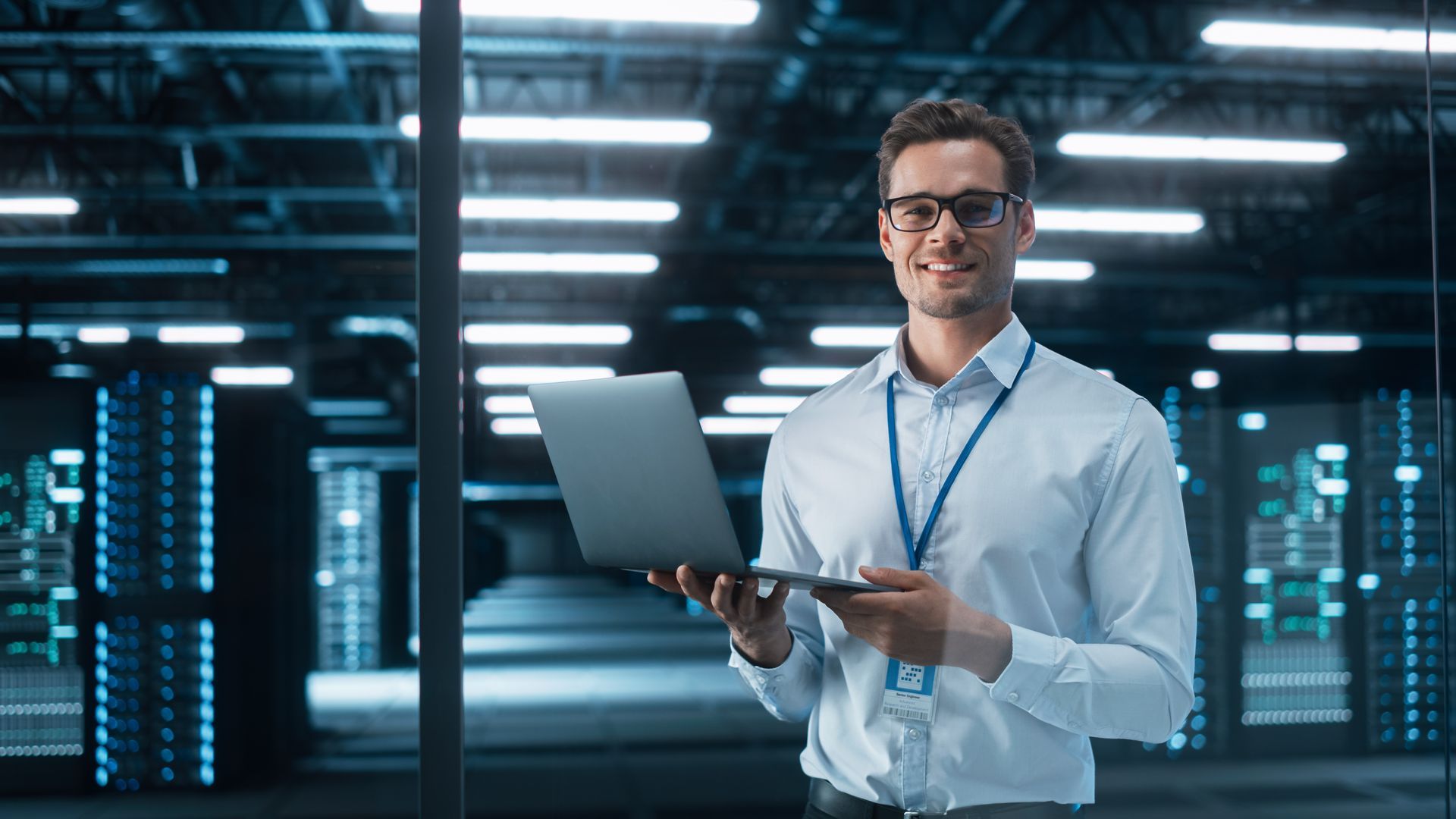 Man with glasses and laptop smiles in a data center, surrounded by servers and lights.