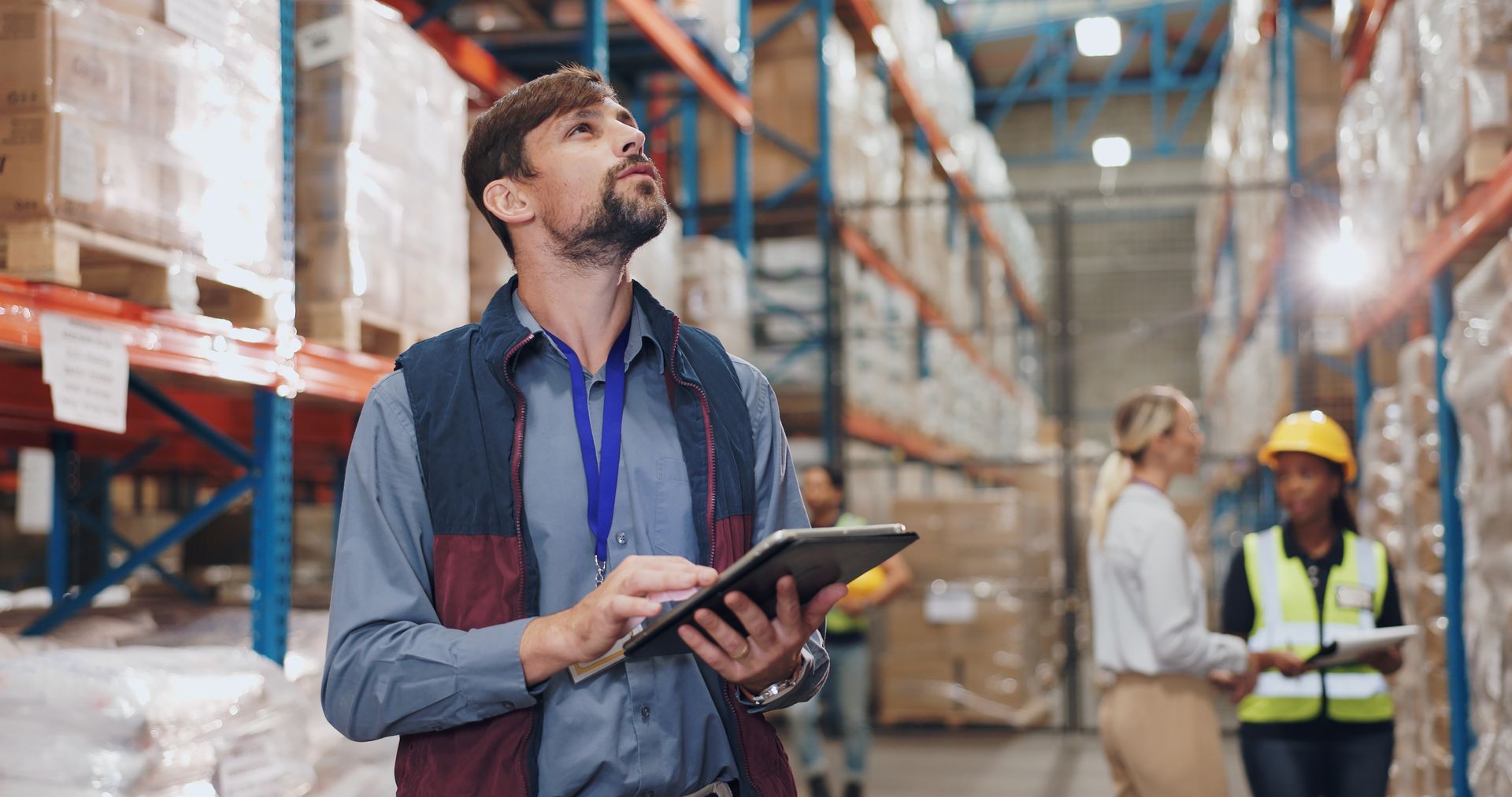 Man with tablet in warehouse, looking up at shelves. Other workers visible in background.