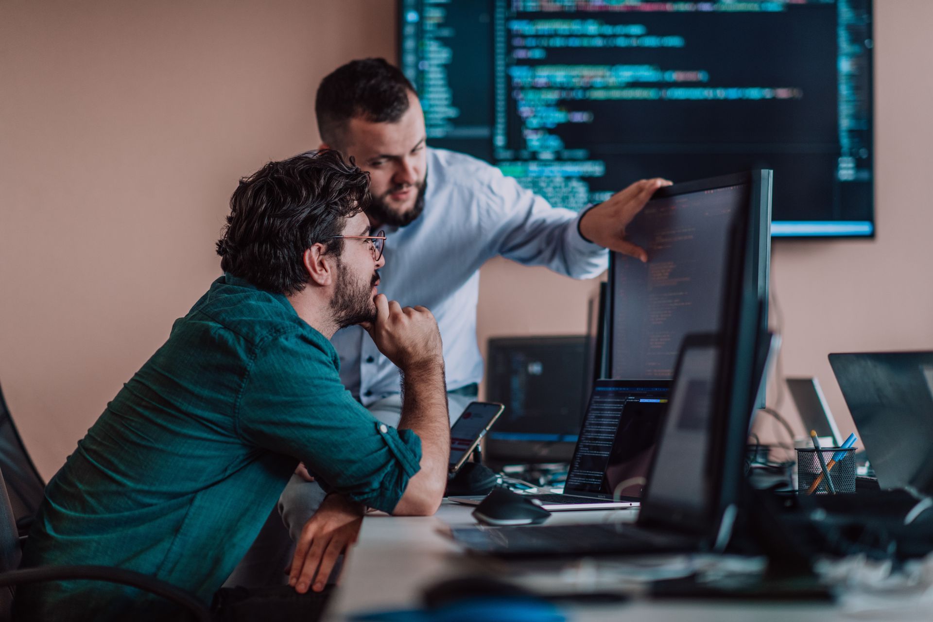 Two men looking at a computer screen, one pointing. Monitors and coding visible; office setting.