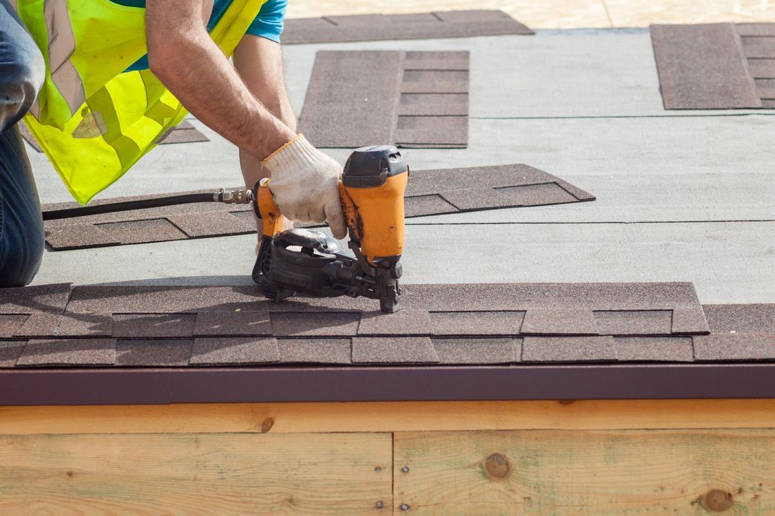 Worker in a yellow vest using a nail gun to install roof shingles on a wooden roof edge