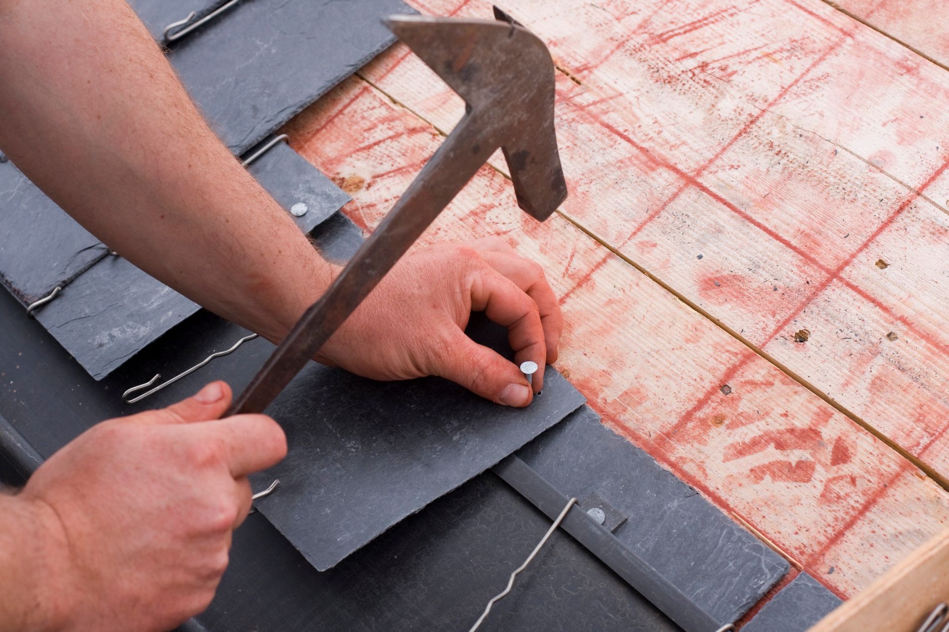 Hands installing black roof shingles with a hammer on a roof surface