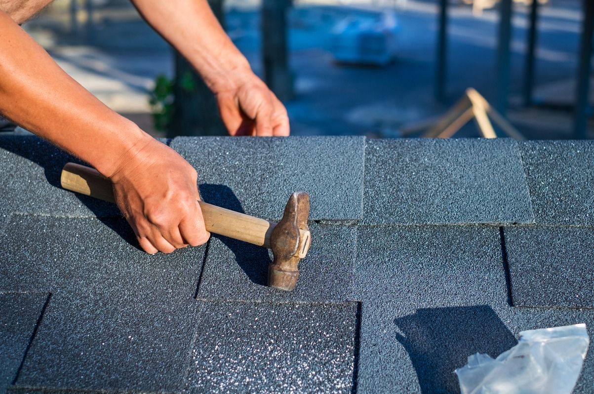 Hands using a hammer to install blue roof shingles on a rooftop.