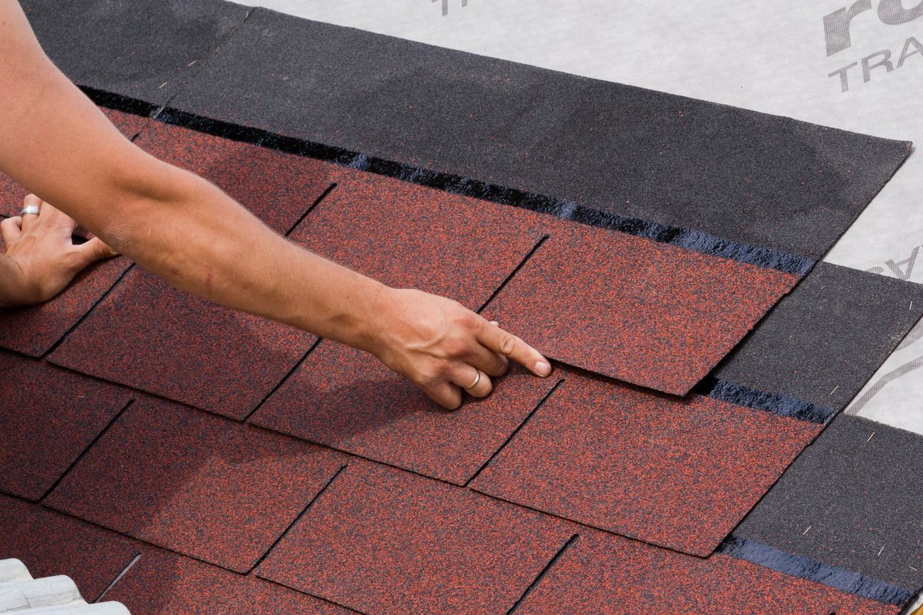 Hands install red roof shingles on a sloped rooftop underlayment.