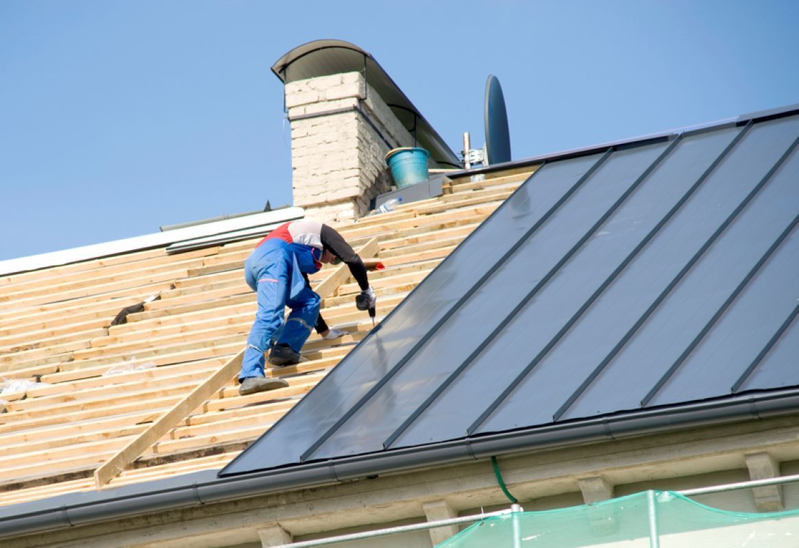 Roofers installing metal roofing on a house roof, one in blue overalls kneeling on plywood sheathing.
