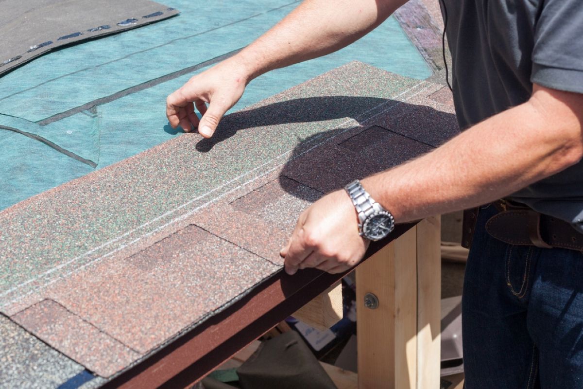 Person installing brown roof shingles on a roof edge with a hand tool