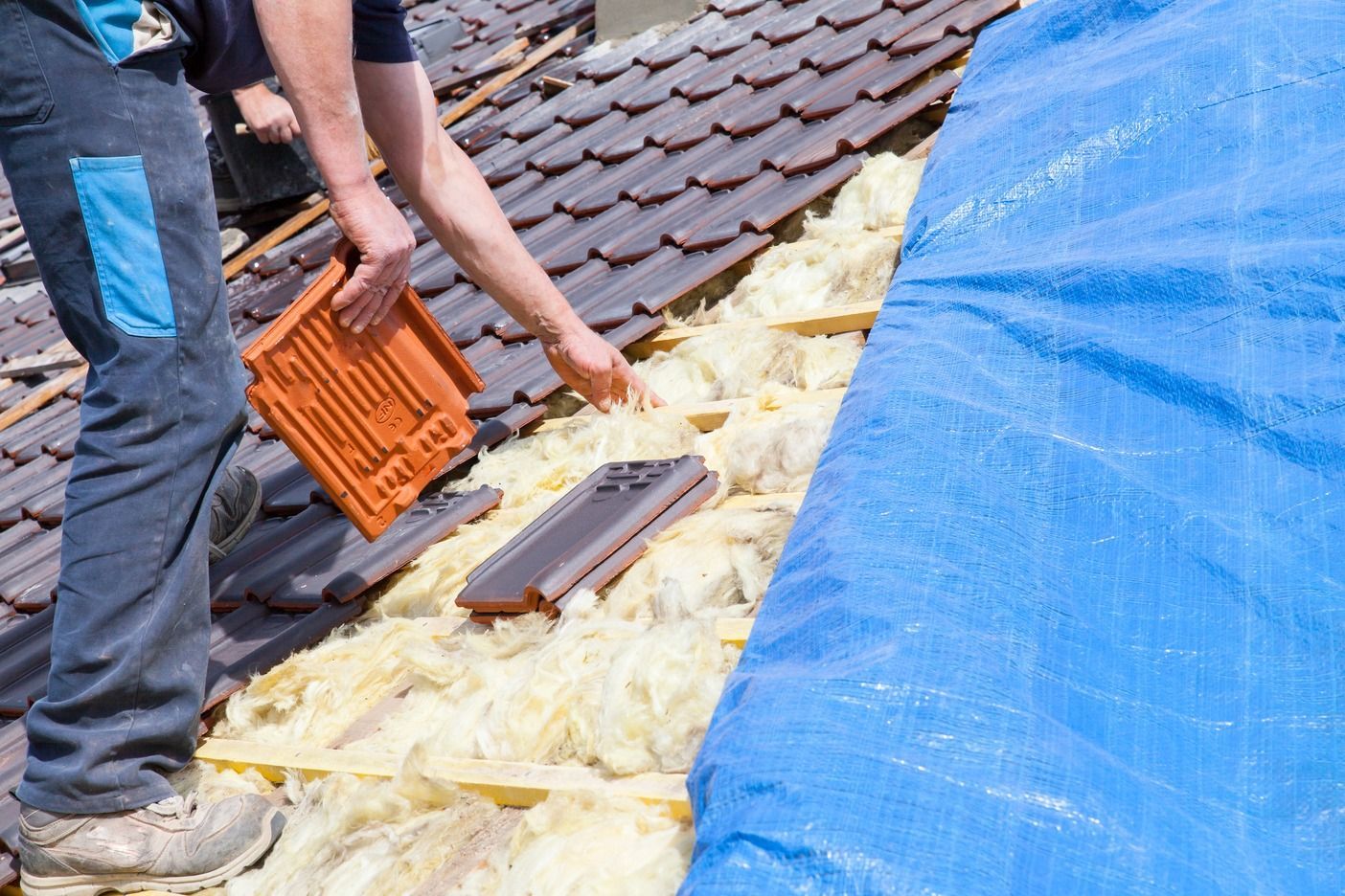 Person installing roof tiles on a house with yellow insulation and a blue tarp covering part of the roof.
