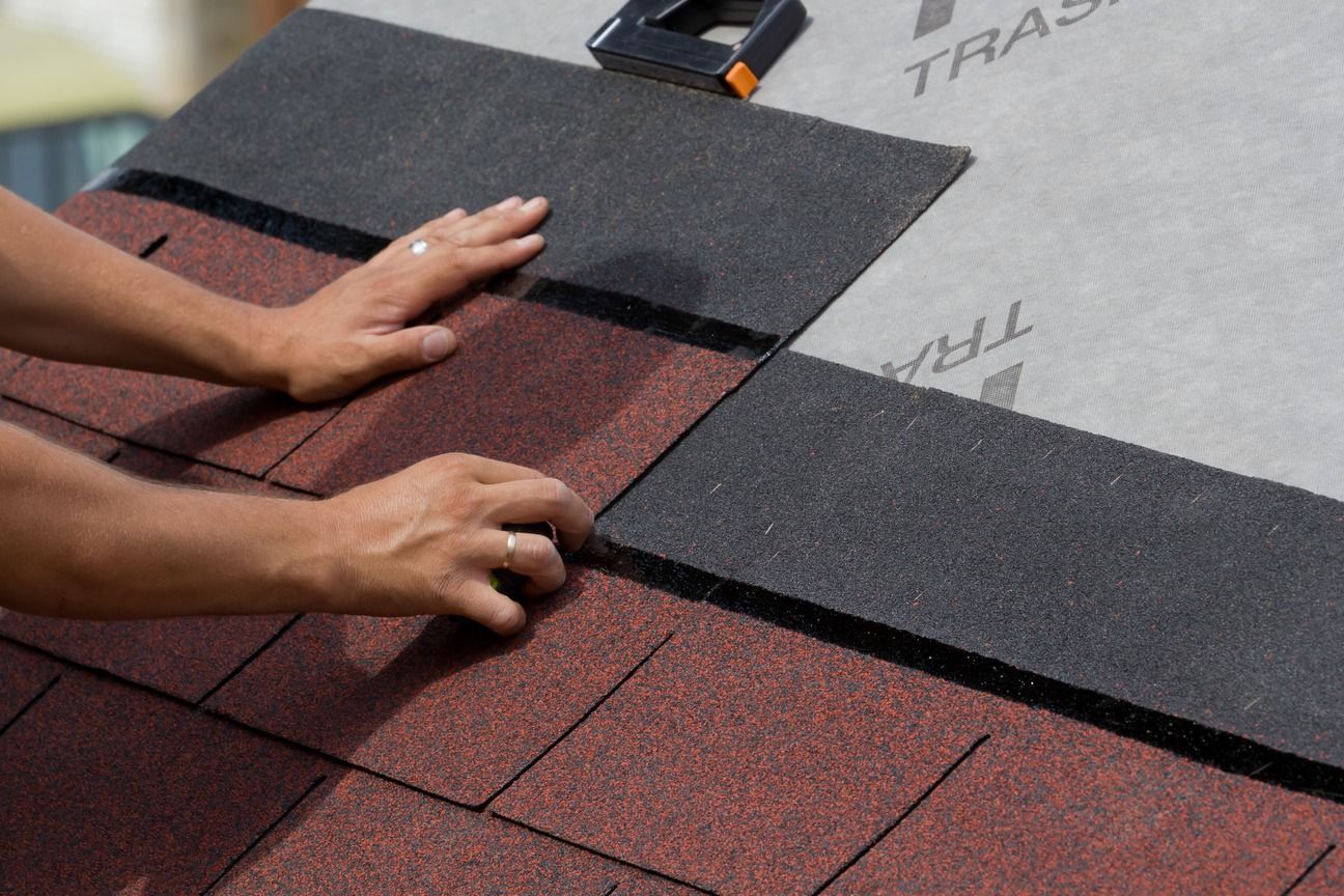 Hands laying black roofing shingles over red shingles on a roof.