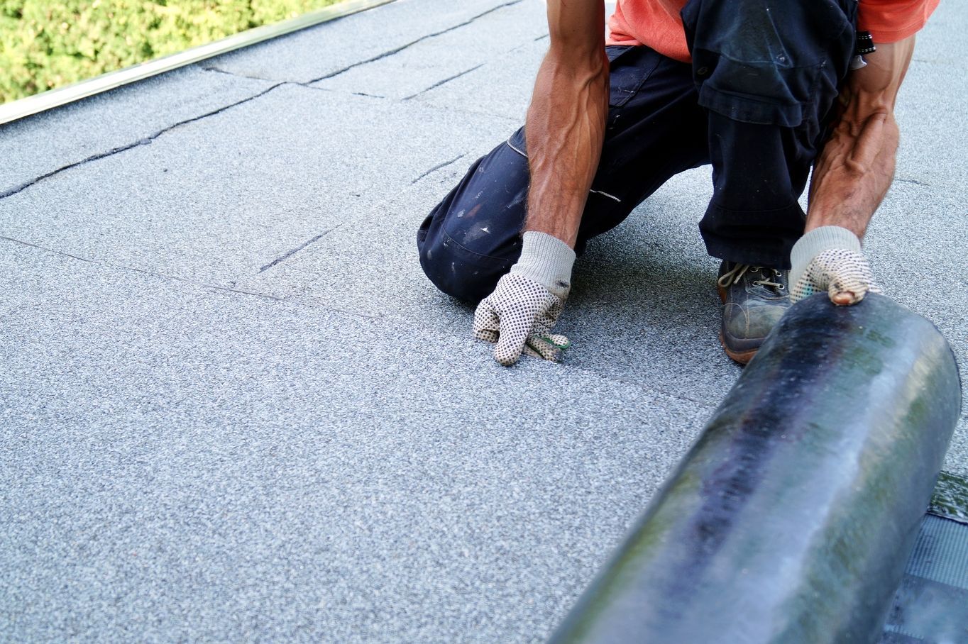 Worker kneeling on a roof, rolling black roofing material over a gray surface.