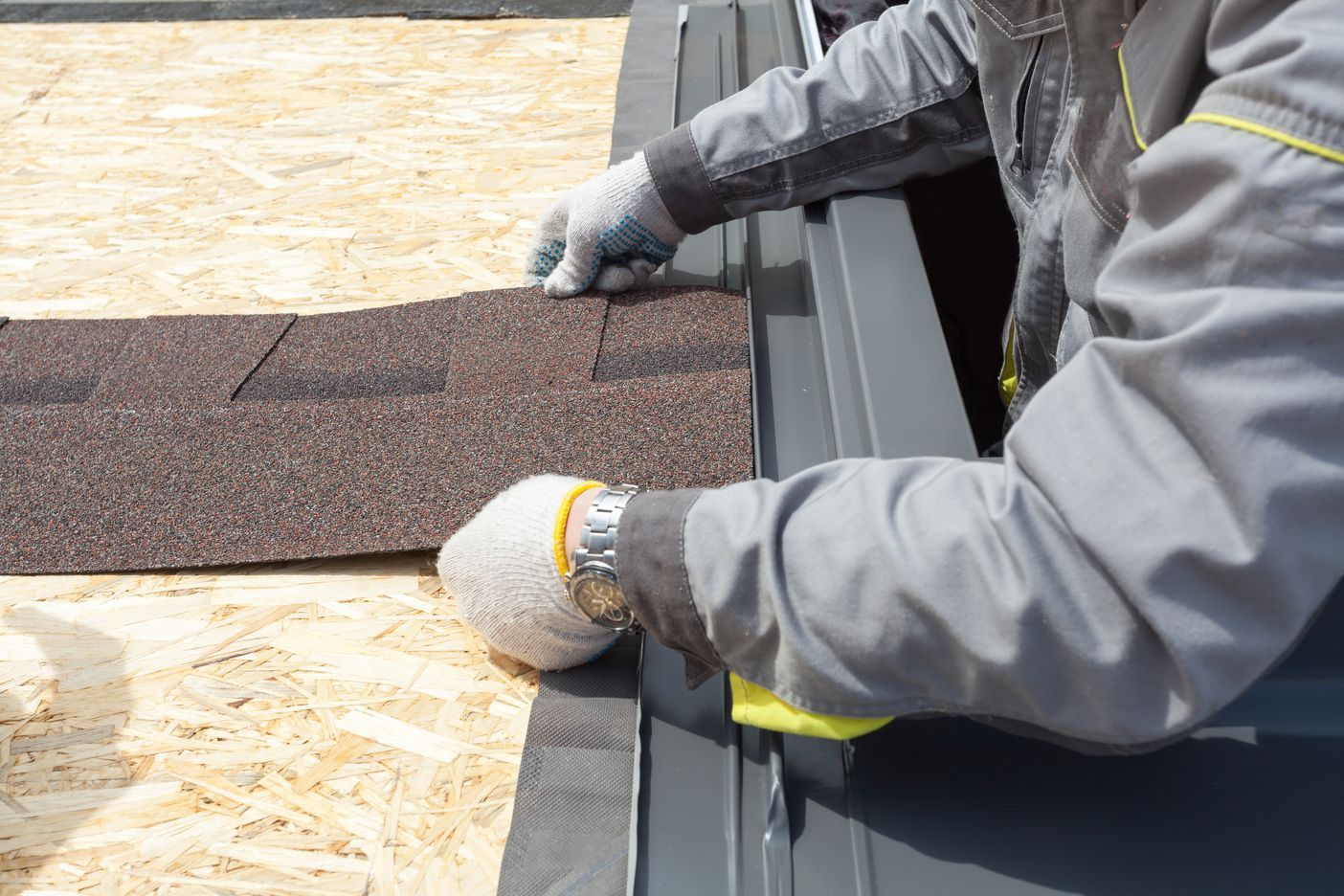 Gloved worker installing brown roof shingles beside gray metal roofing edge