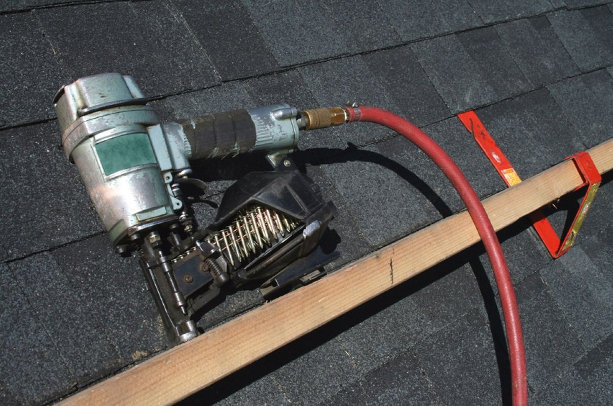 Roofing nail gun connected to a red air hose on dark shingles, with a wooden handle in the foreground