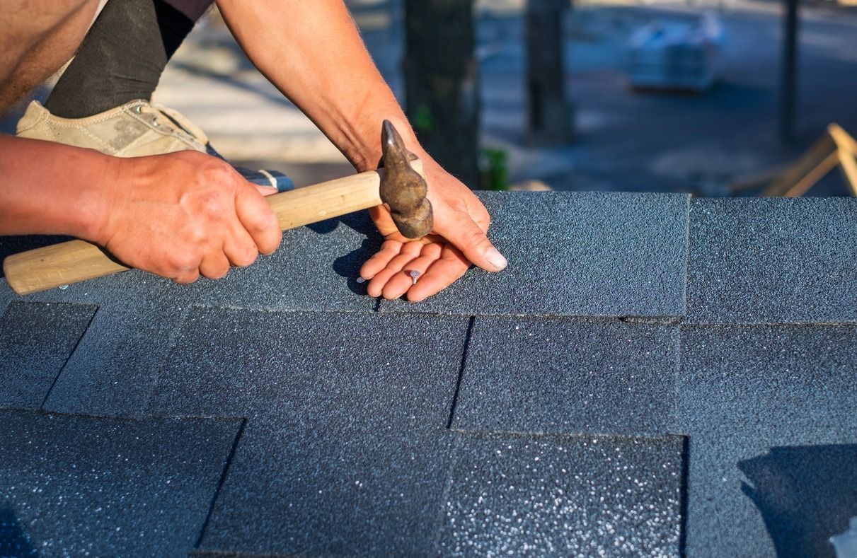Hands hammering roofing shingles on a blue-gray roof surface