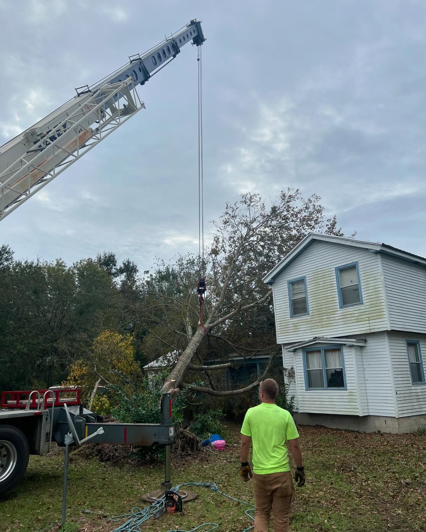 Fire truck crane lifts a tree branch beside a damaged white house; person in neon shirt watches.