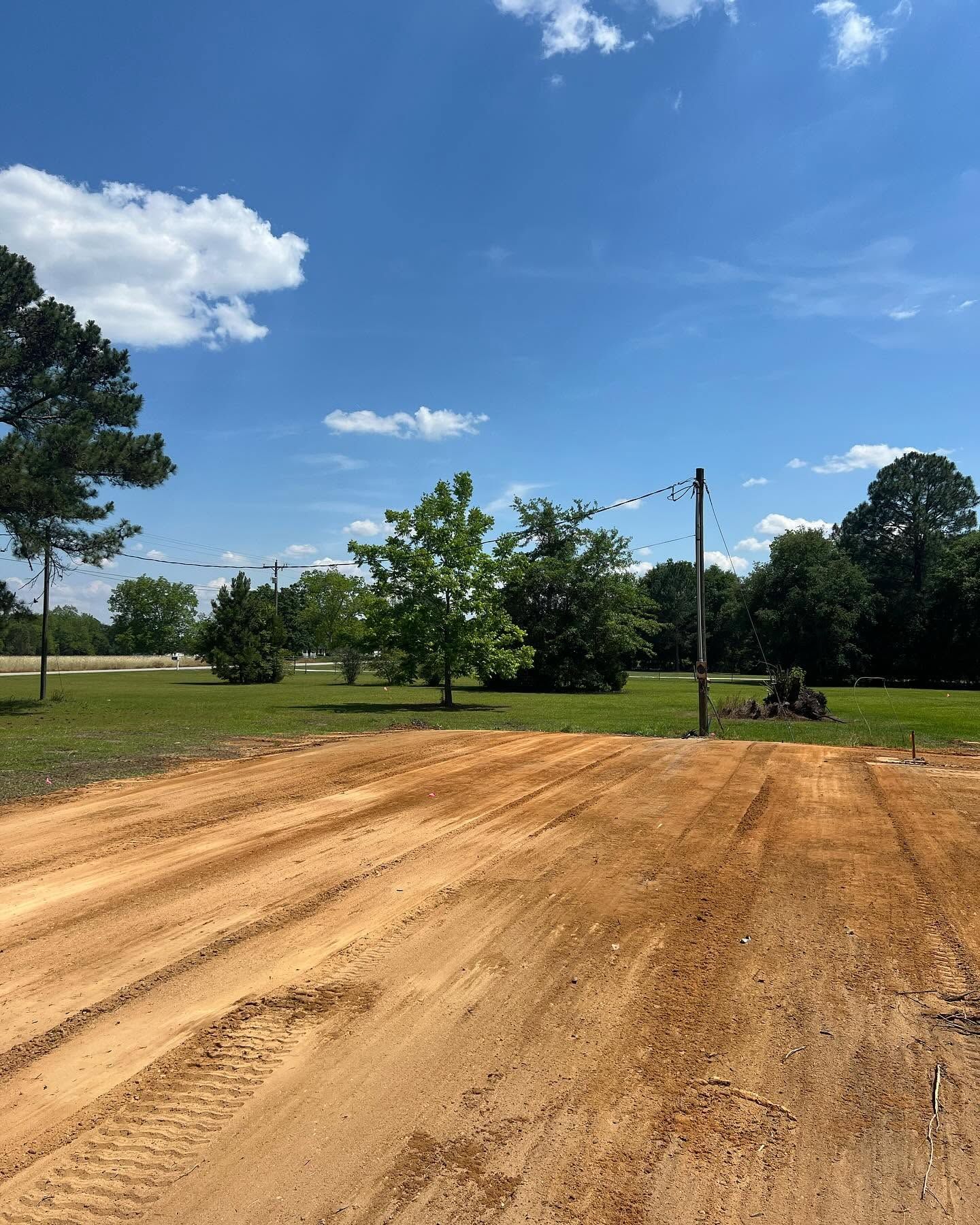 Freshly graded dirt lot under a bright blue sky with trees and utility poles in the distance
