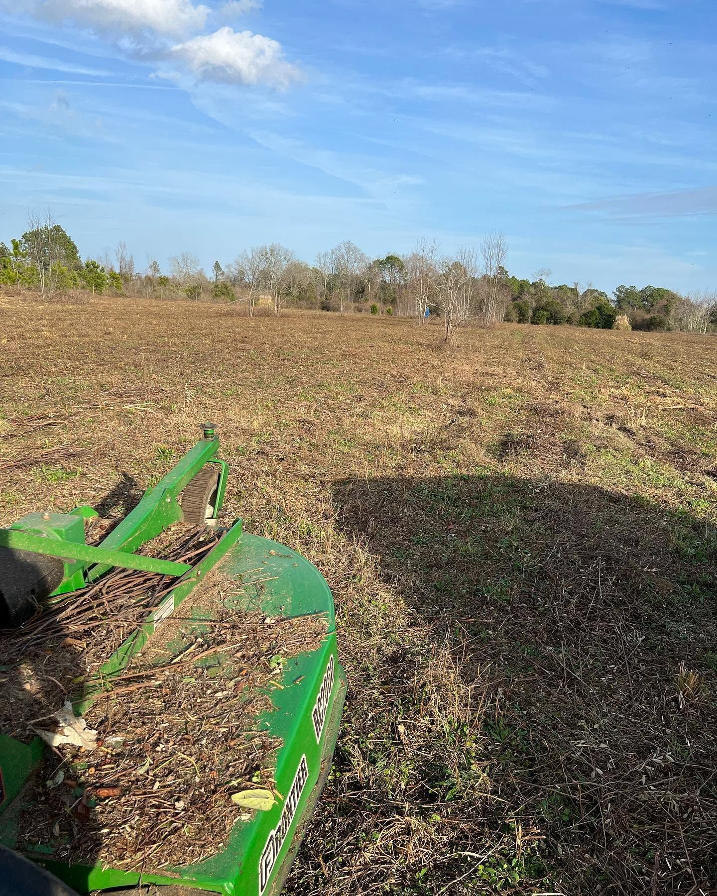 Green farm implement in a harvested field under a blue sky, with trees along the horizon.