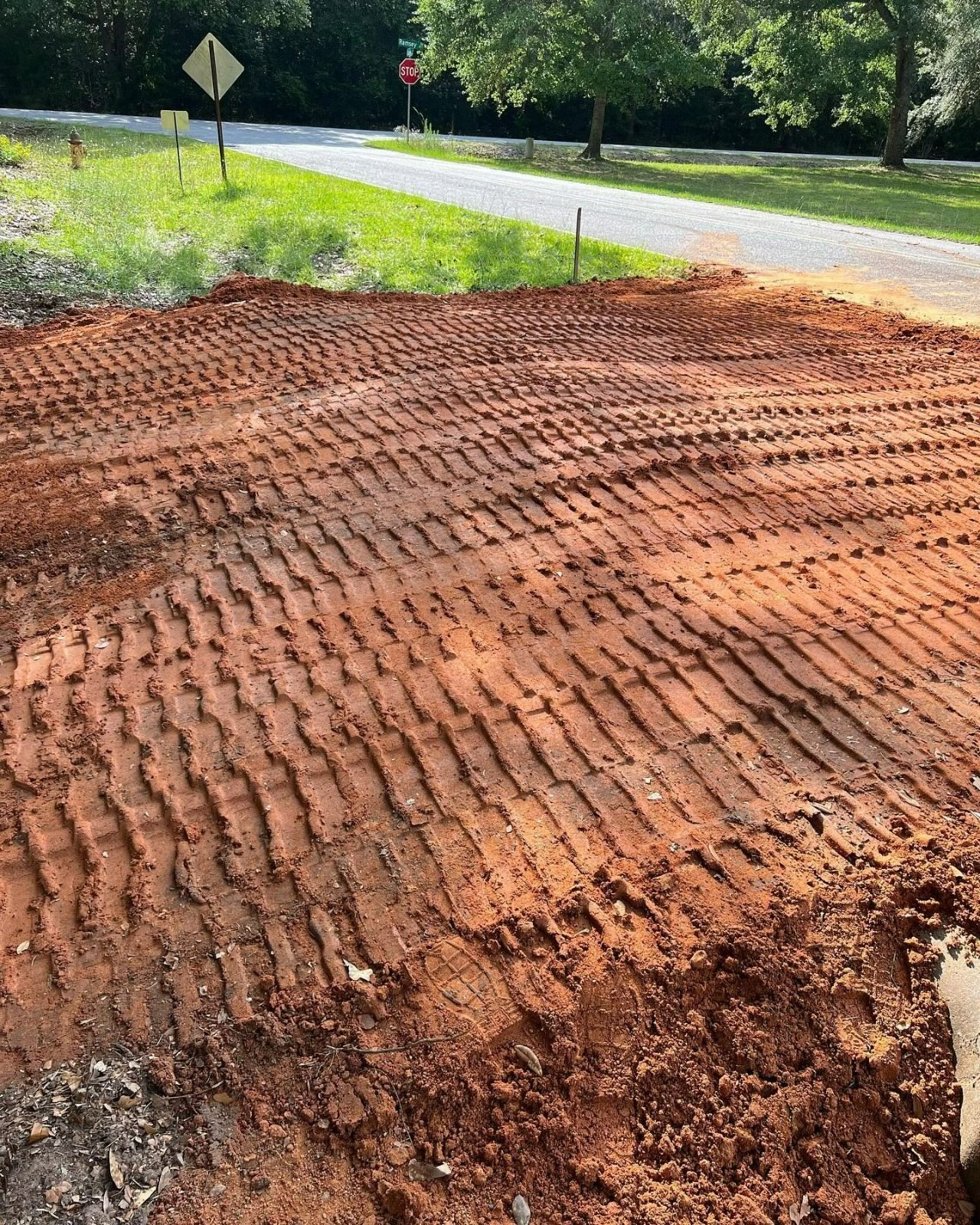 Freshly graded dirt beside a road, with tire tracks and a grassy roadside in the background