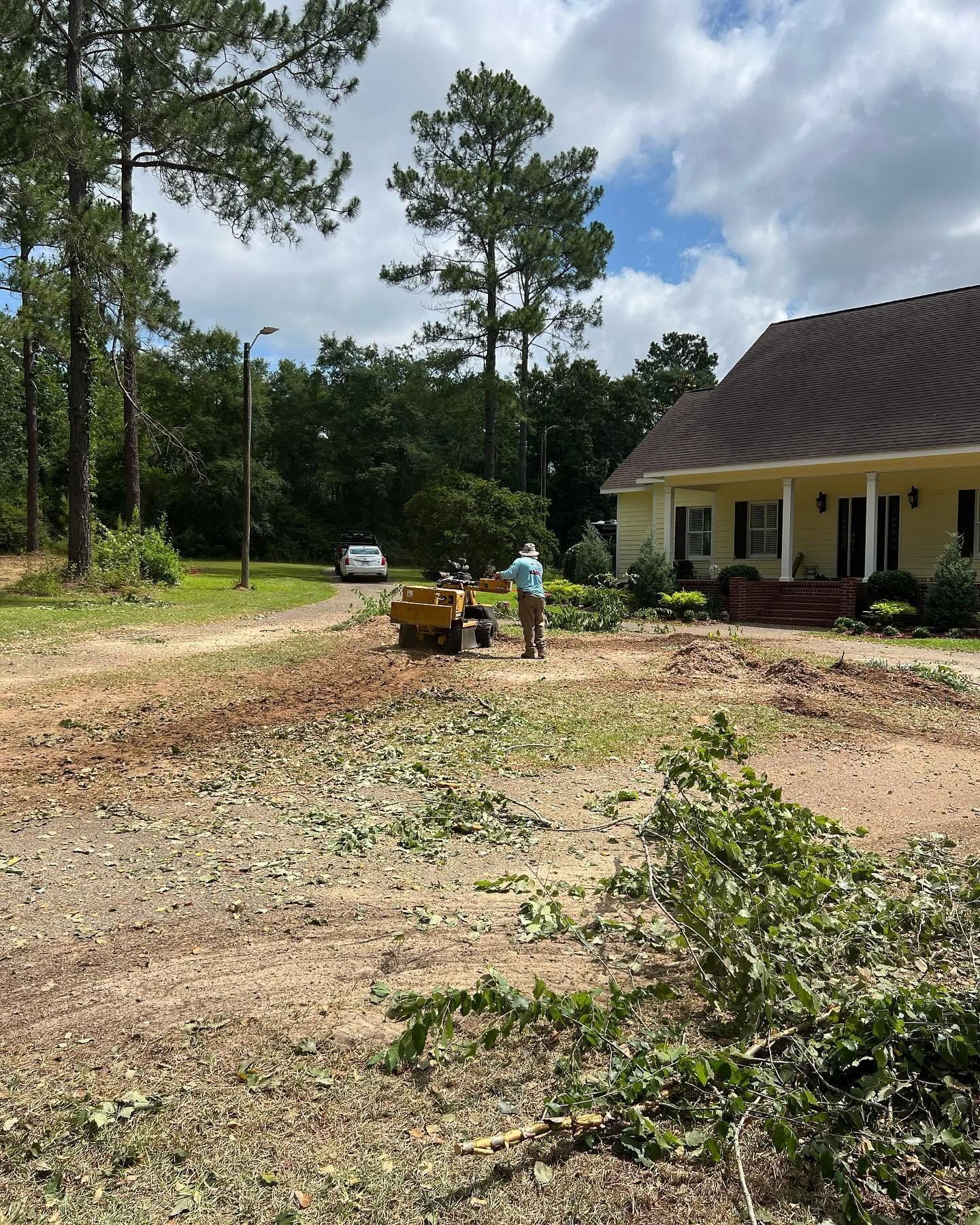 Person moving debris in a yard beside a yellow house, with trees and a parked car nearby.