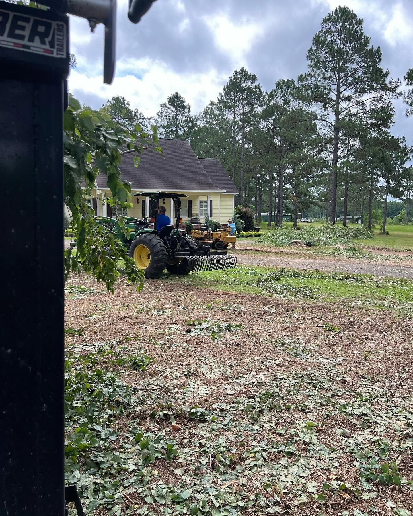 Person on a tractor in a yard beside a house and pine trees under a cloudy sky