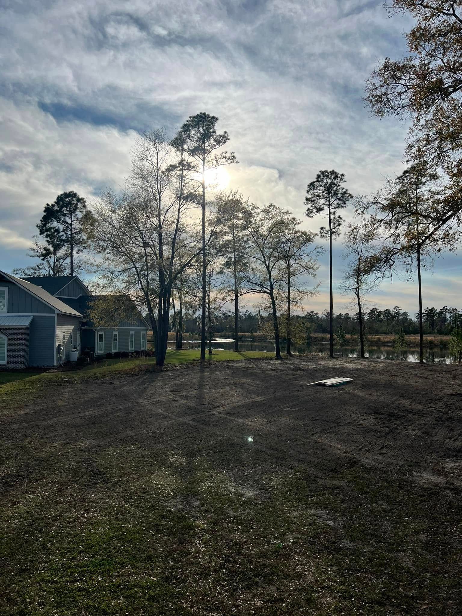 Rural yard with bare trees, a house on the left, and the sun glowing through clouds over open fields