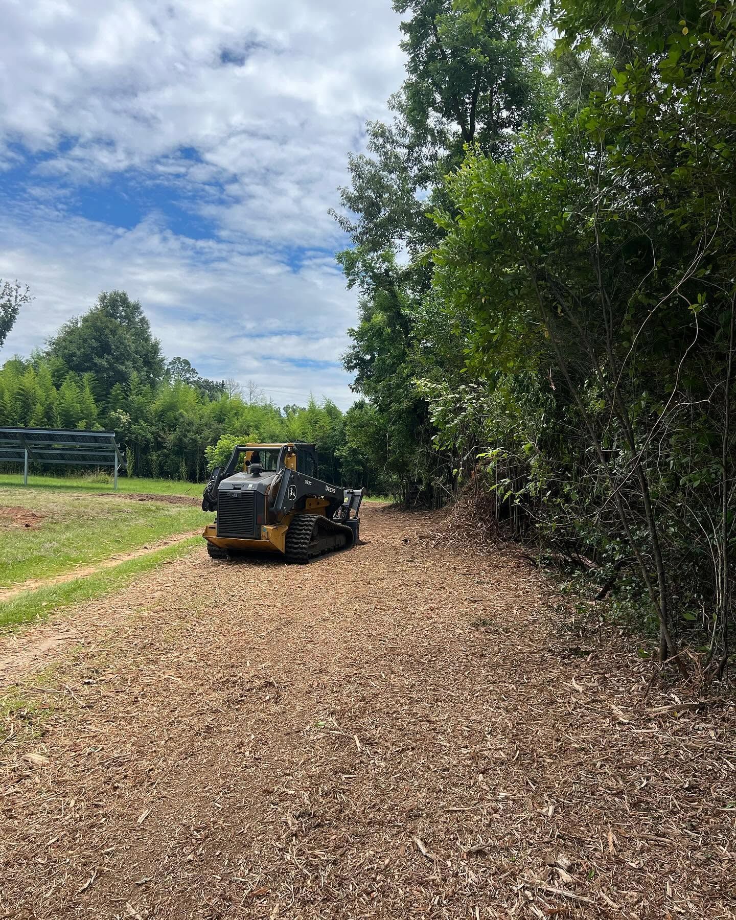 Small black skid steer clearing a woodchip path beside trees under a partly cloudy sky