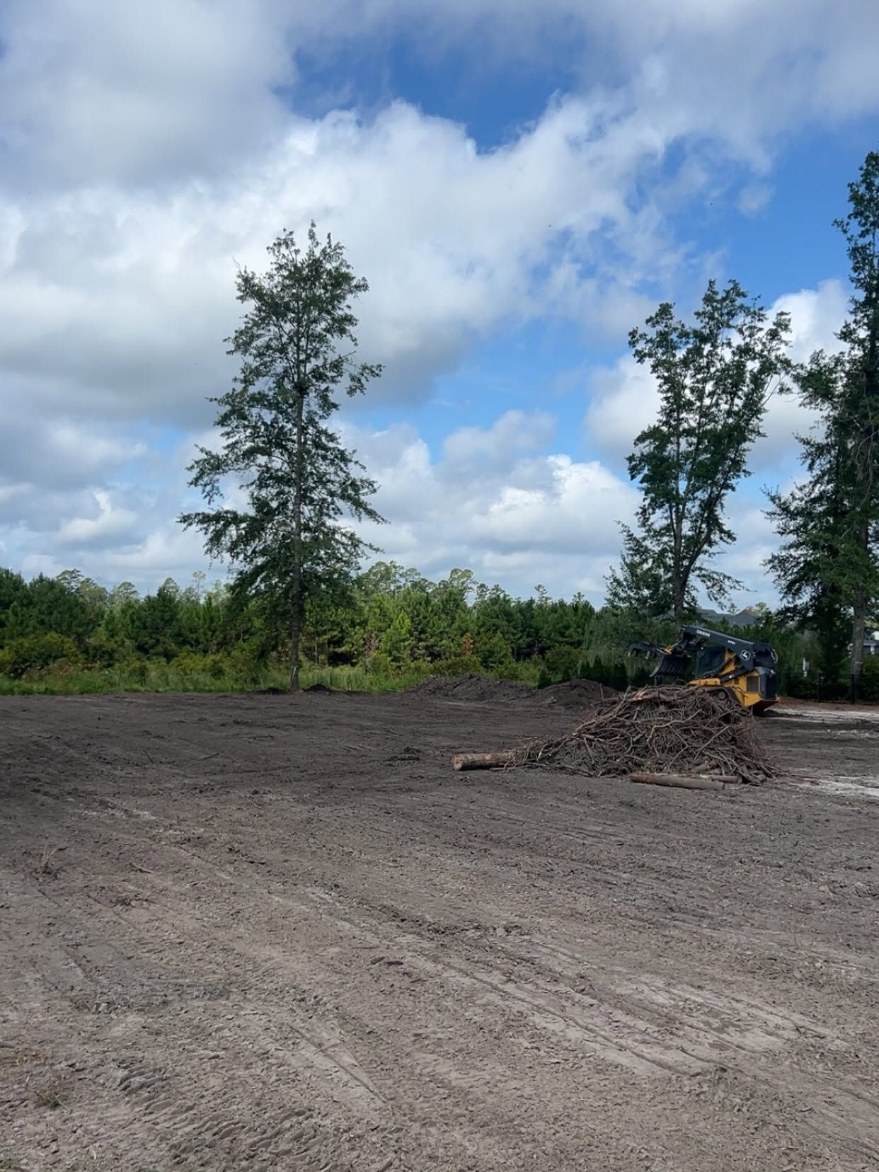 Cleared dirt lot under a cloudy sky, with trees and a small yellow excavator in the distance