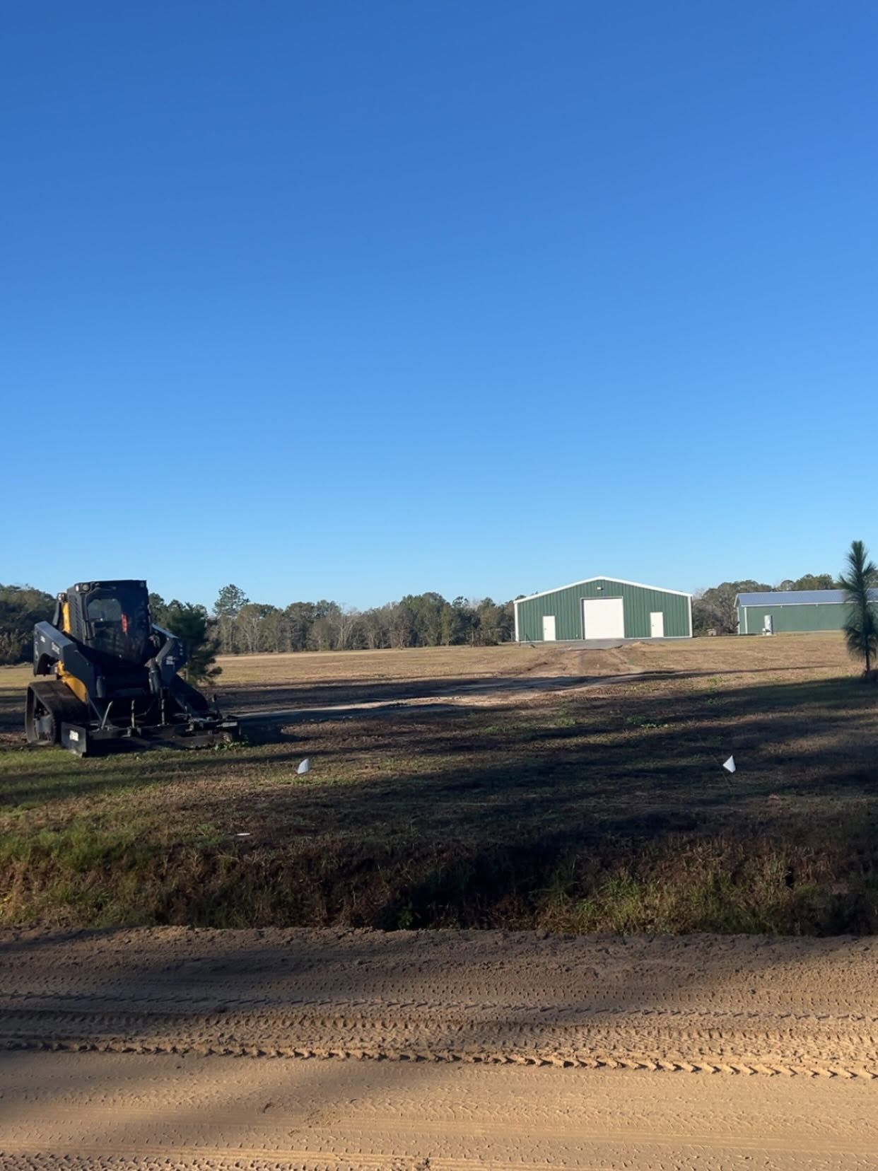 Construction equipment on a dirt lot near a green barn under a clear blue sky