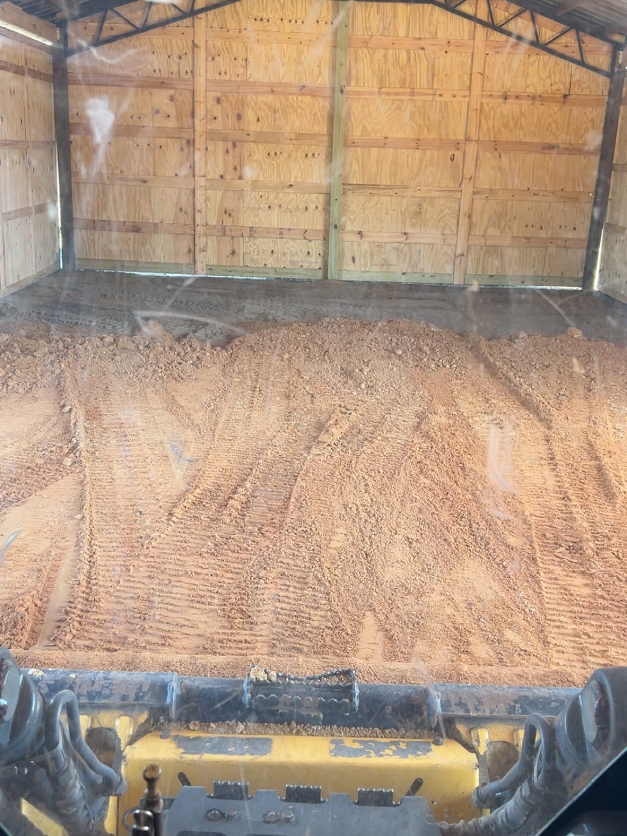 Inside a barn, a yellow skid steer pushes a large pile of tan grain or silage.