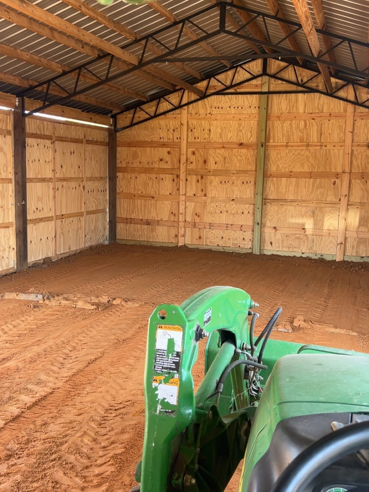Inside a wooden barn, a green tractor faces a dirt floor under a metal roof.