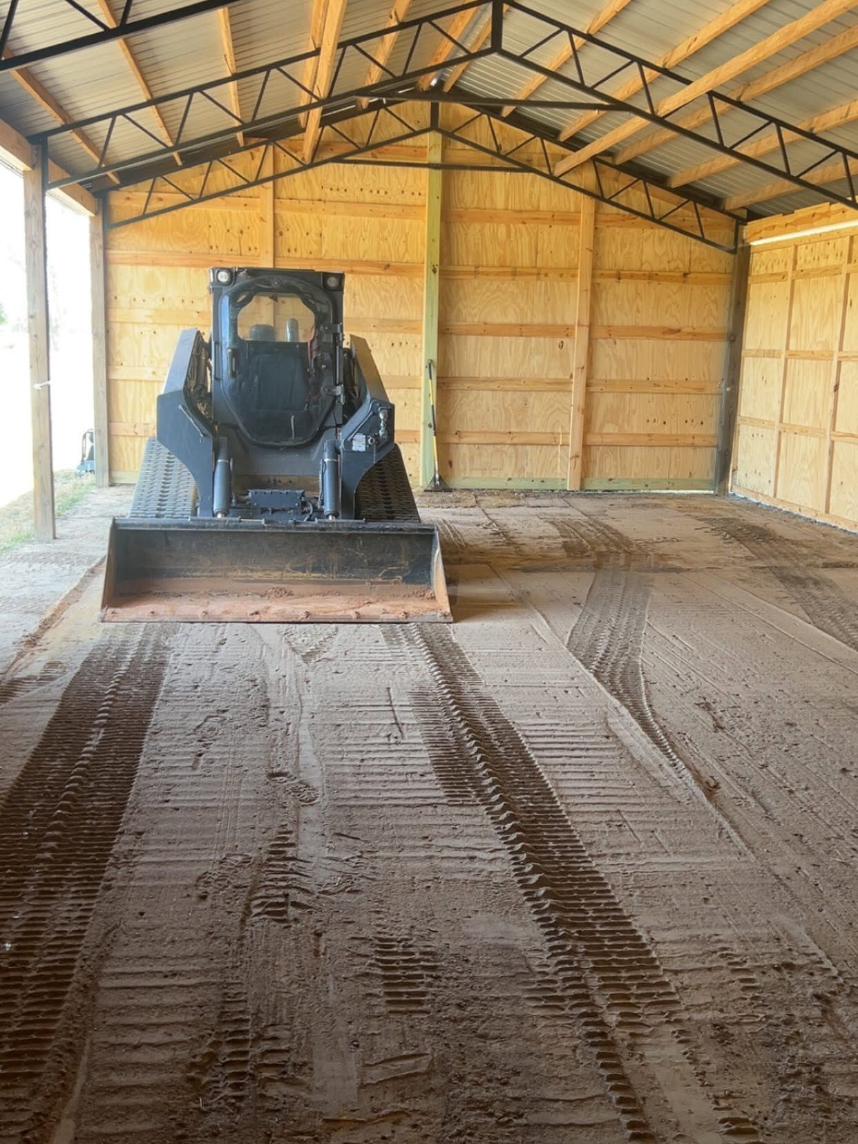 Skid steer in a wooden barn, leveling dirt across the floor under a metal roof.