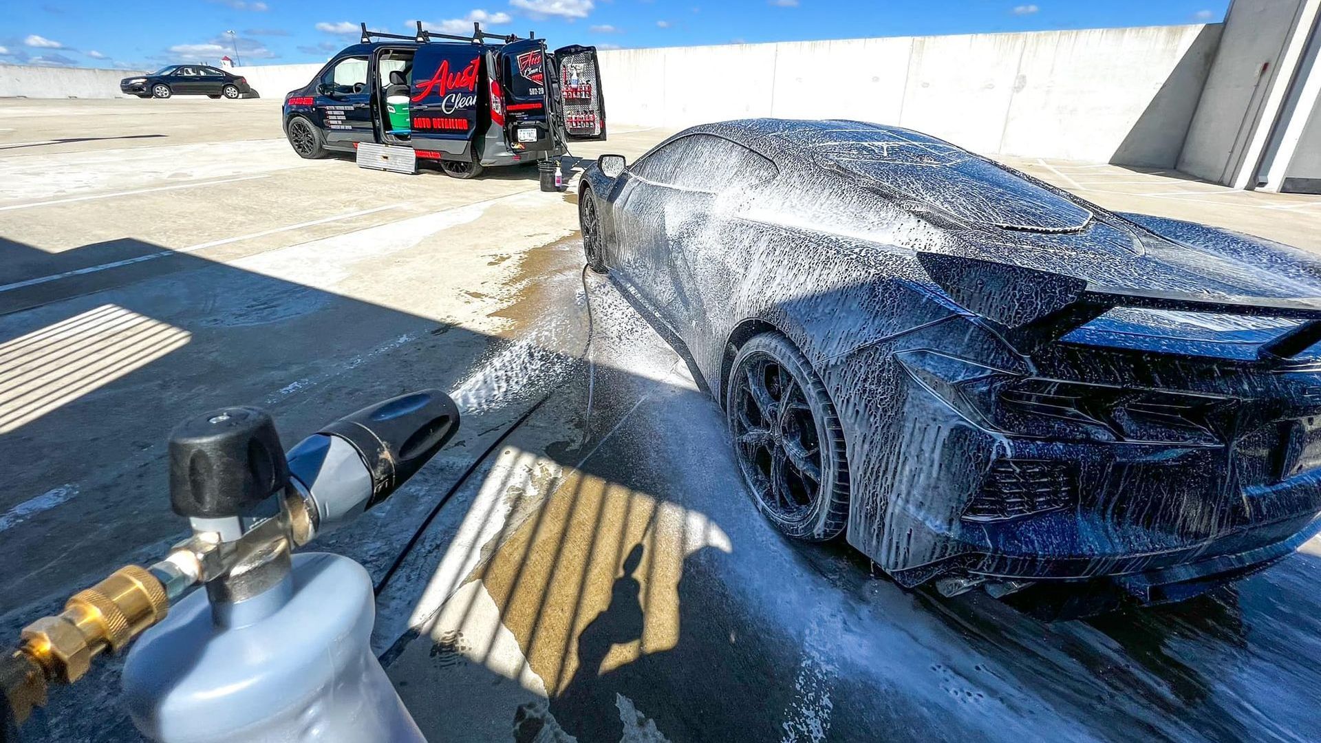 A black sports car covered in foam is being washed outdoors near a parked van and building.