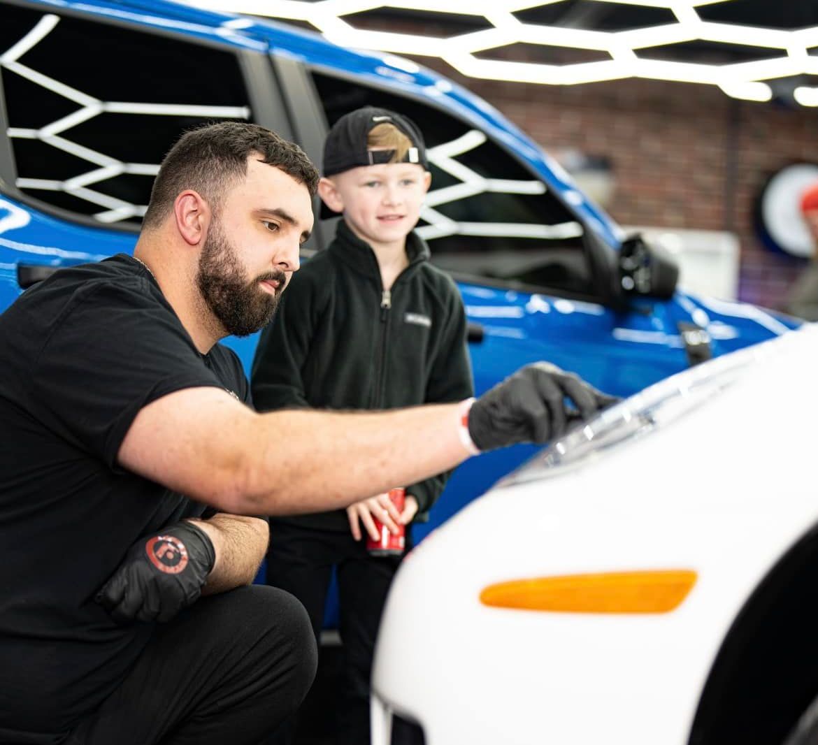 Man and boy examining a car headlight in a detailing shop. Man points, boy smiles.