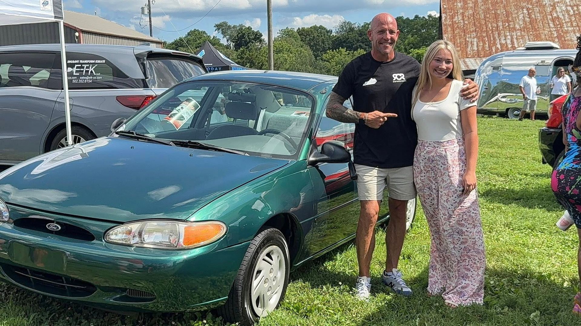Man points at a green Ford Escort with a woman standing next to it in a grassy outdoor setting.