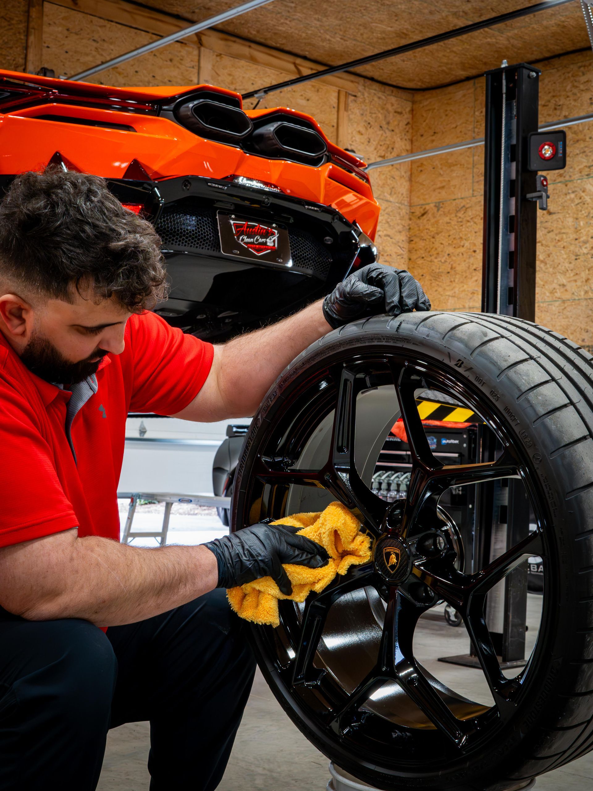 Man in red shirt cleans a black car rim with a yellow cloth. Orange car is in background.