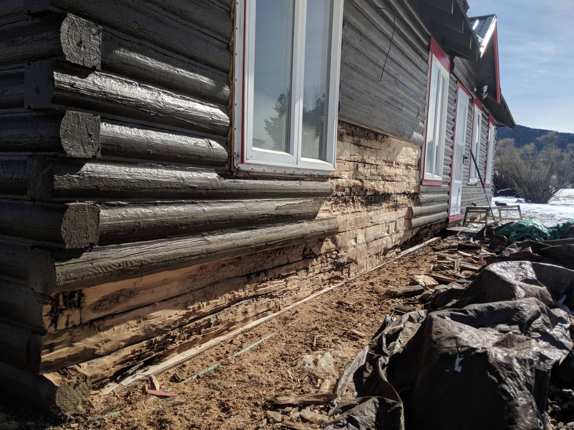Log cabin wall with severe erosion along the foundation in a muddy yard near snow and mountains.