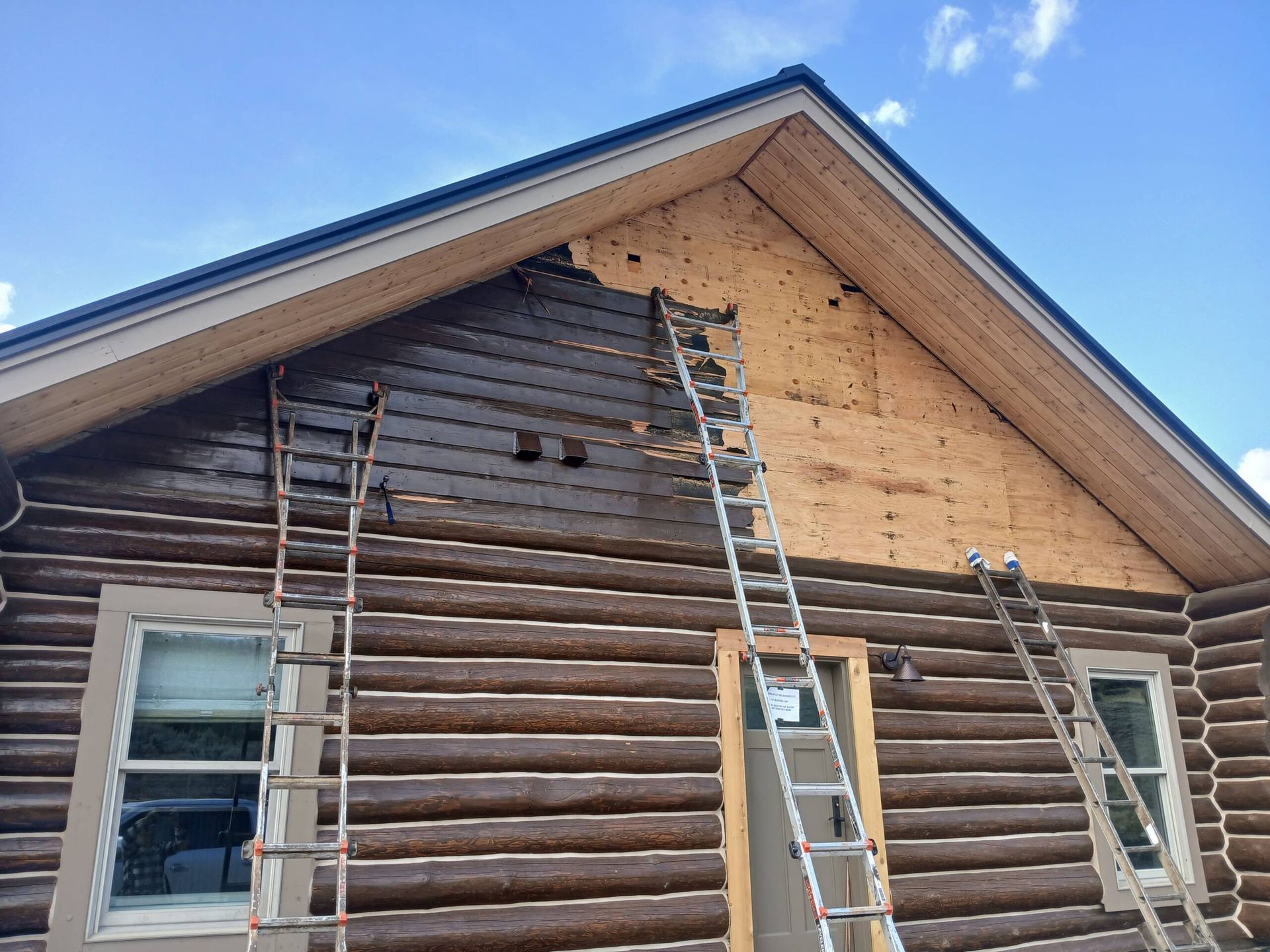 House under repair with exposed log siding, ladders leaning against the wall, and bare plywood on the gable end