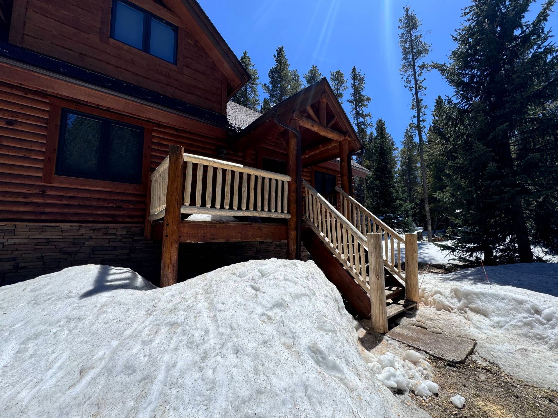 Log cabin with wooden porch and stairs in a snowy forest under a clear blue sky