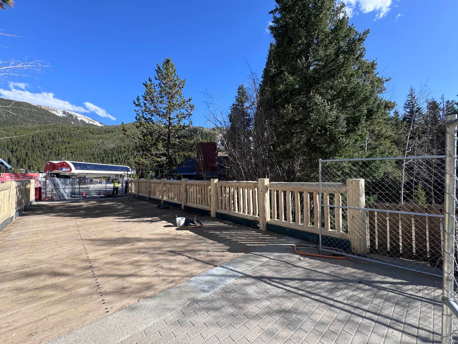 Wooden fence along a gravel driveway beside trees and mountains under a clear blue sky