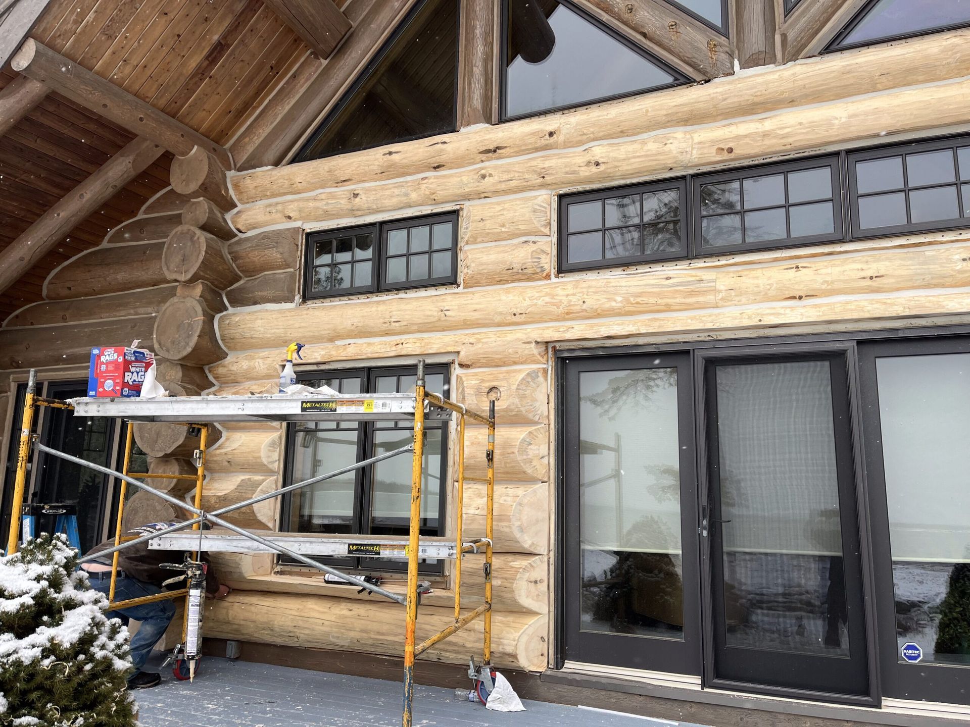Scaffold beside a log cabin exterior with black-framed windows and sliding glass doors in a snowy setting