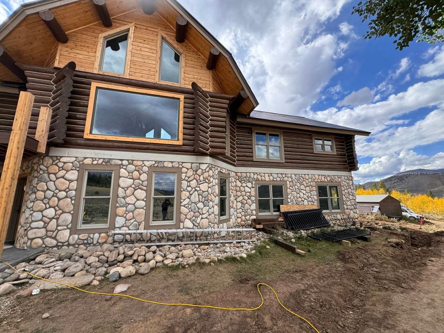 Rustic log cabin with stone foundation under construction on a dirt lot, cloudy sky overhead
