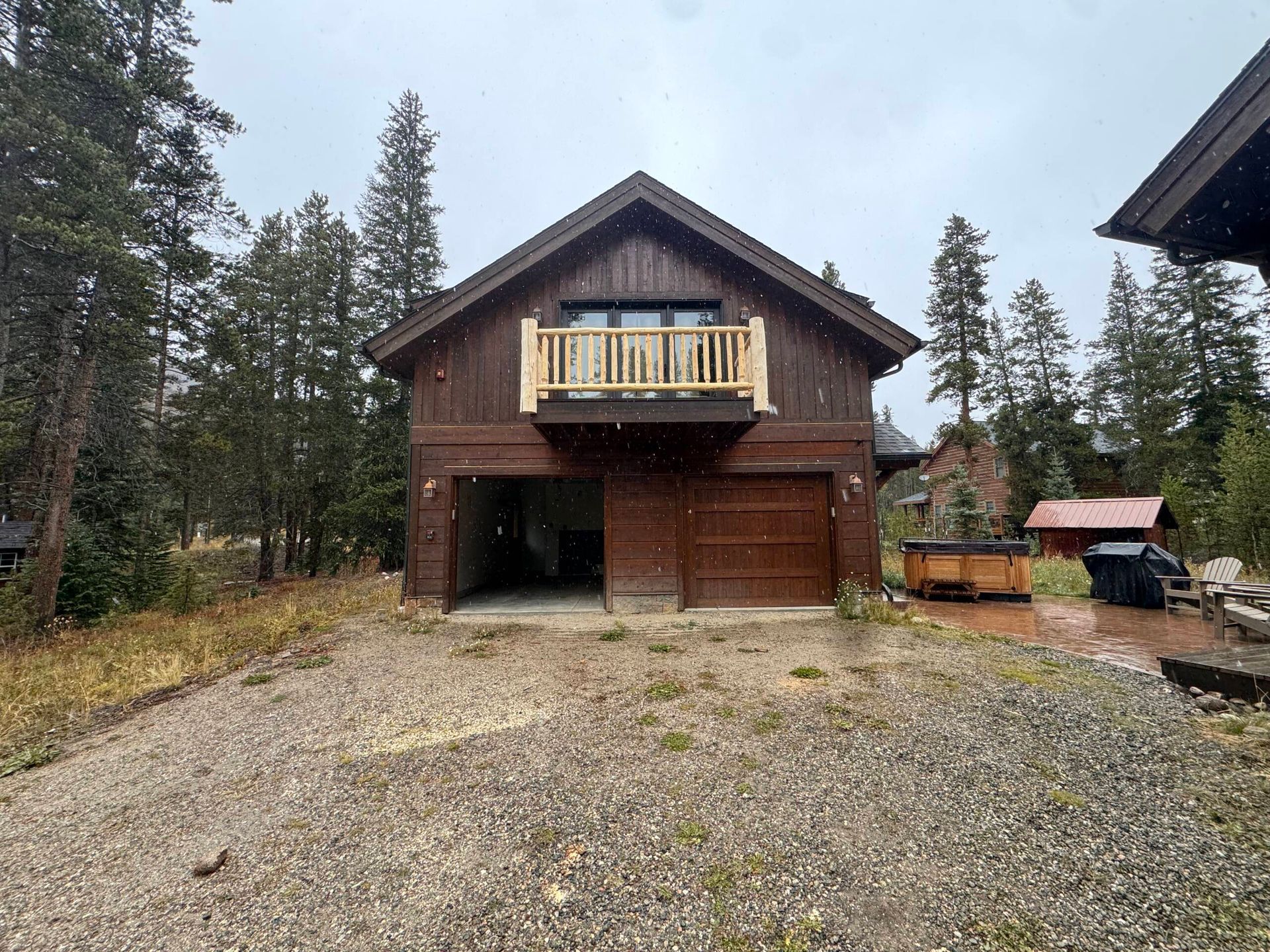 Rustic wooden cabin with balcony and garage, set on a gravel driveway surrounded by trees.