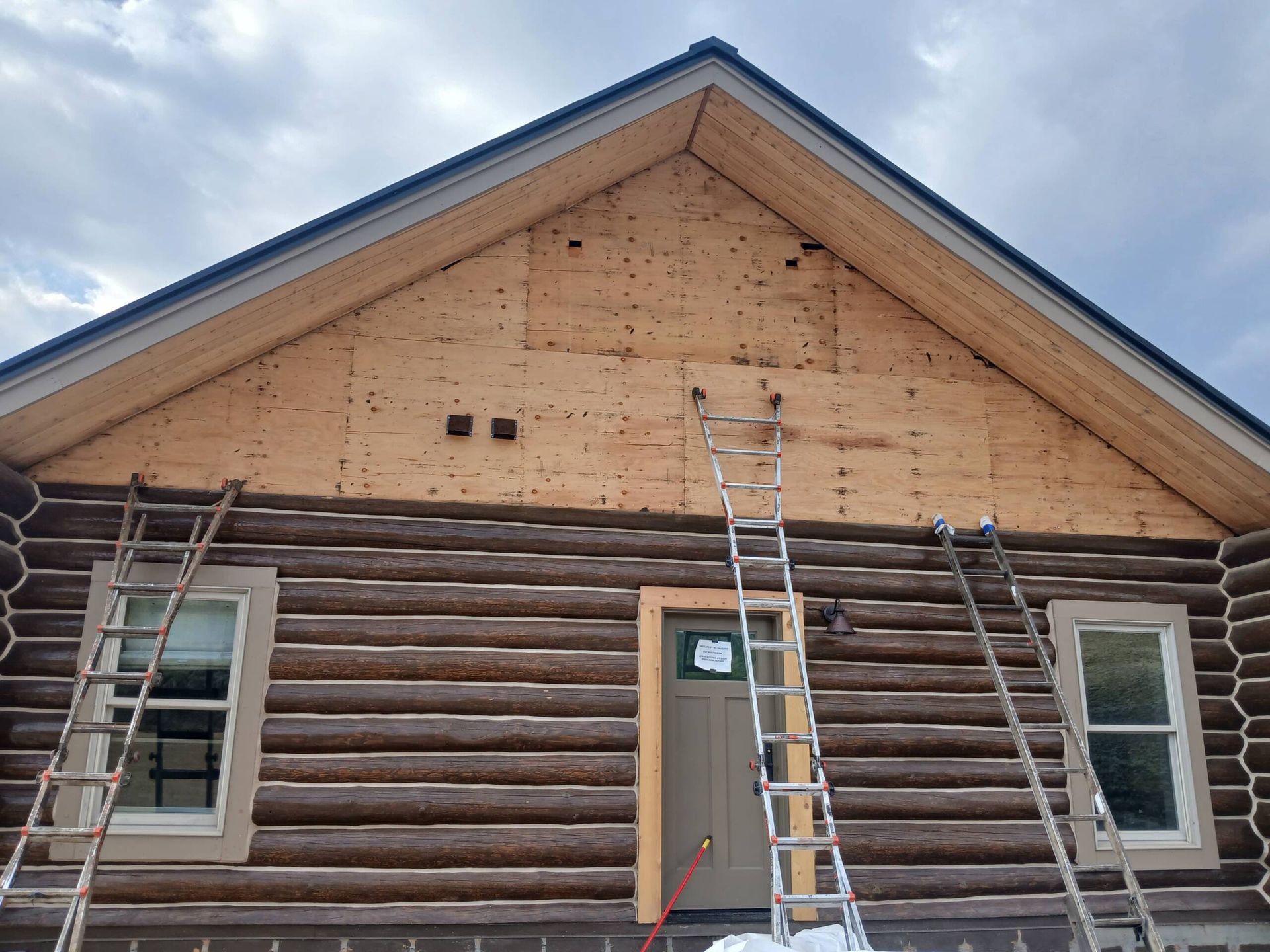 Log cabin under repair with two ladders leaned against the front wall and a small window on each side.
