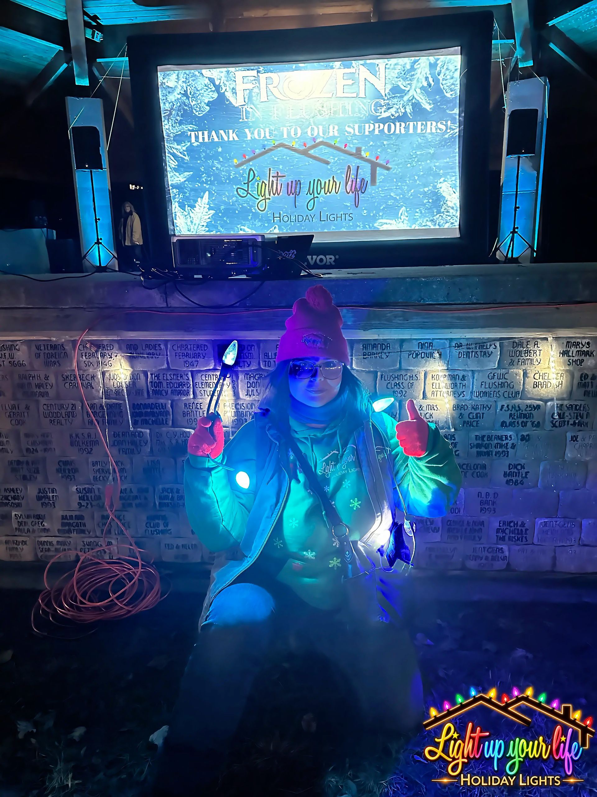 Frozen theme light display in Fenton Michigan. owner Lindsay holding blue and teal lights. blue and teal flood uplighting in background with light up your life holiday lights logo on the big screen. 