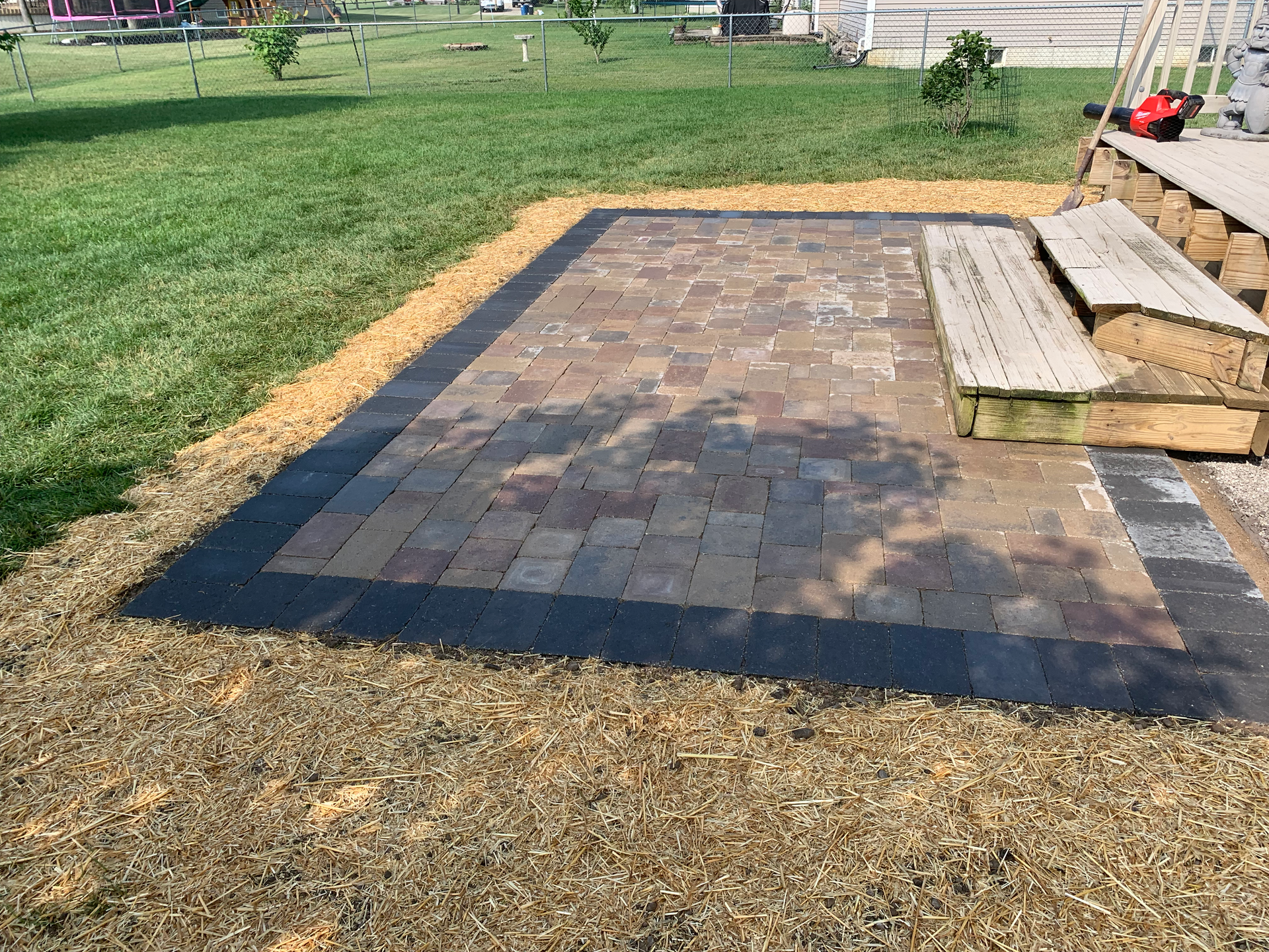 A brick patio with a wooden bench in the middle of a yard.