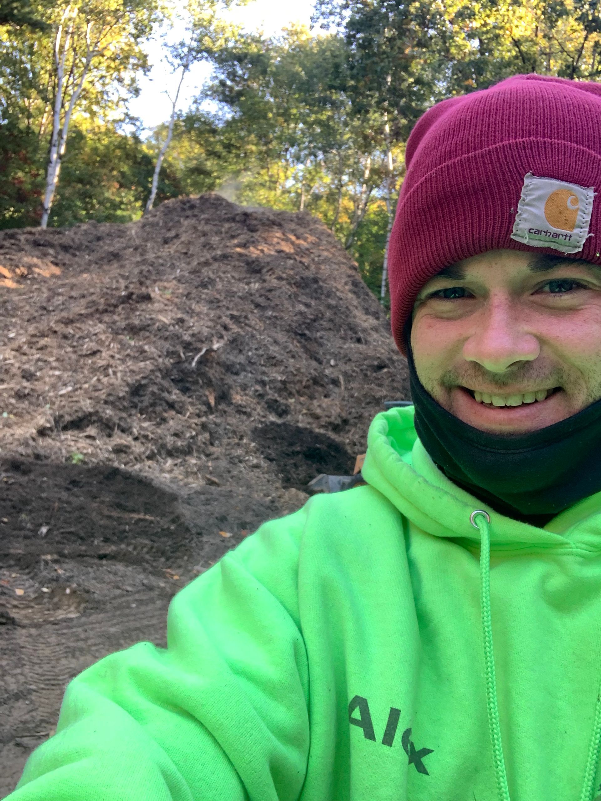 A man wearing a green hoodie and a red beanie is smiling in front of a pile of dirt.
