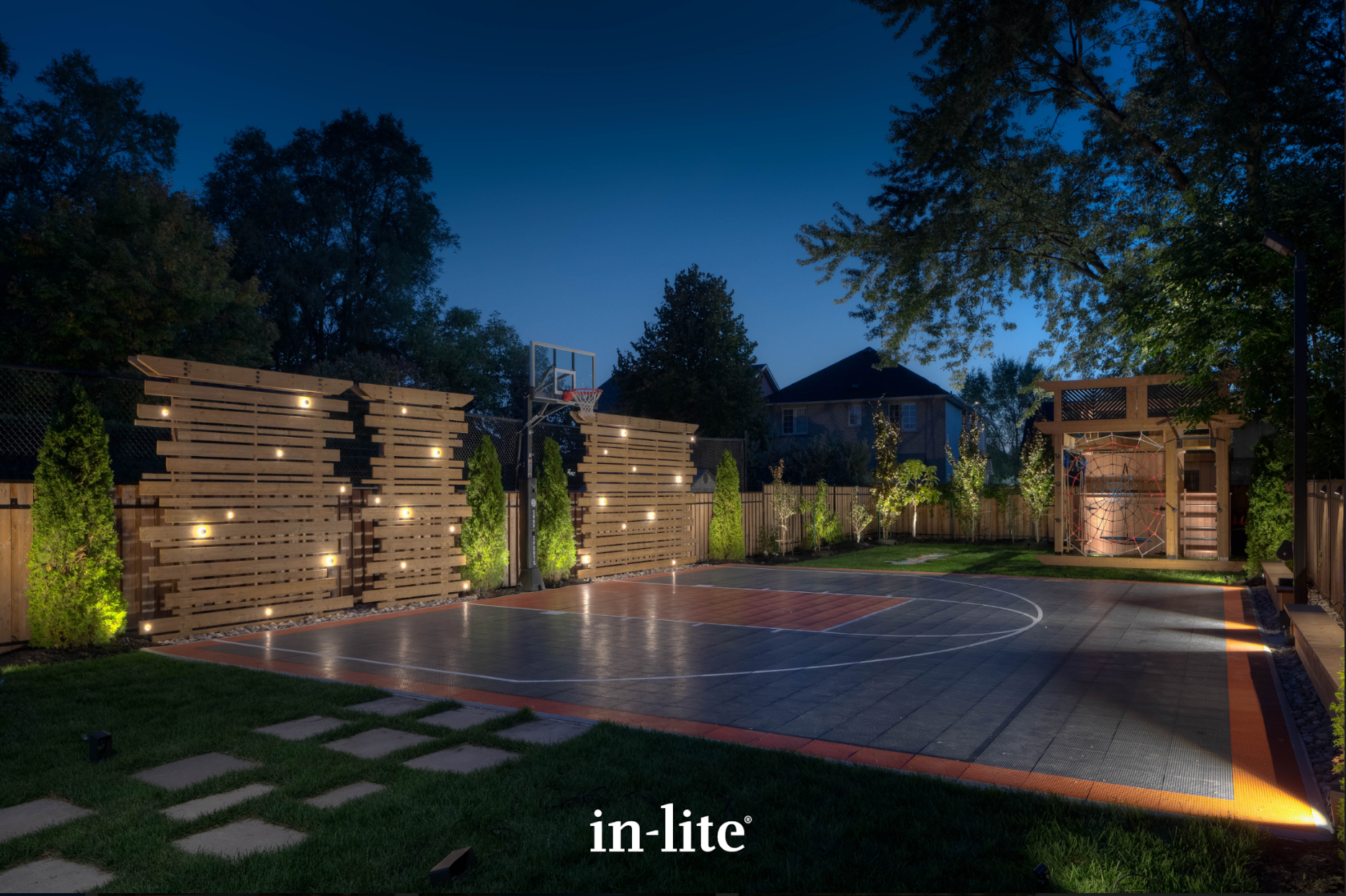 A basketball court is lit up at night in a backyard. landscape lighting.